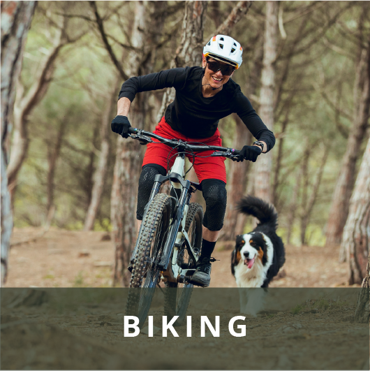 Smiling woman mountain biking with a Bernese Mountain dog following her through a forest.